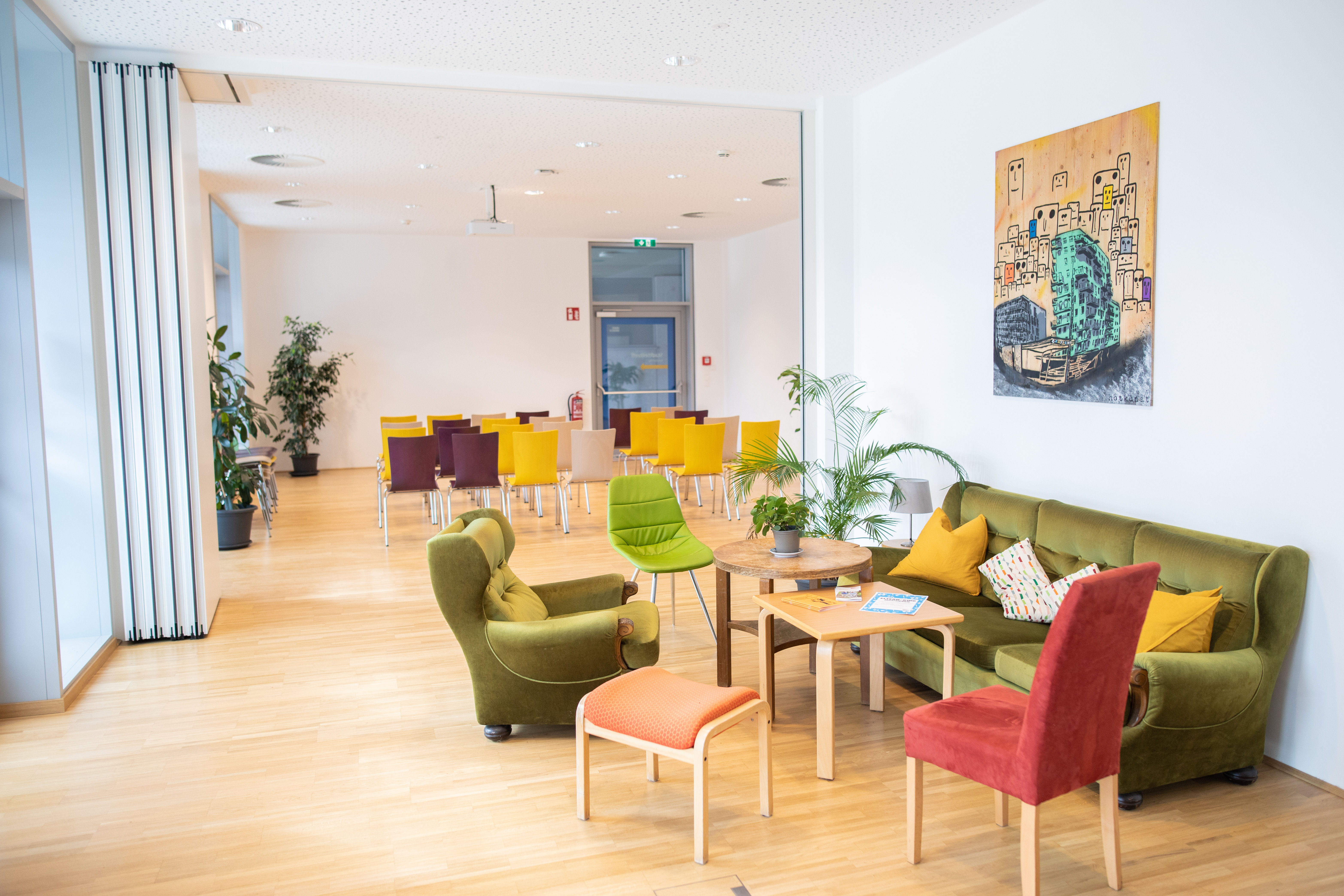 Heller Aufenthaltsraum mit grünen und roten Sofas, einem Tisch und Pflanzen, im Hintergrund ein Essbereich mit bunten Stühlen.