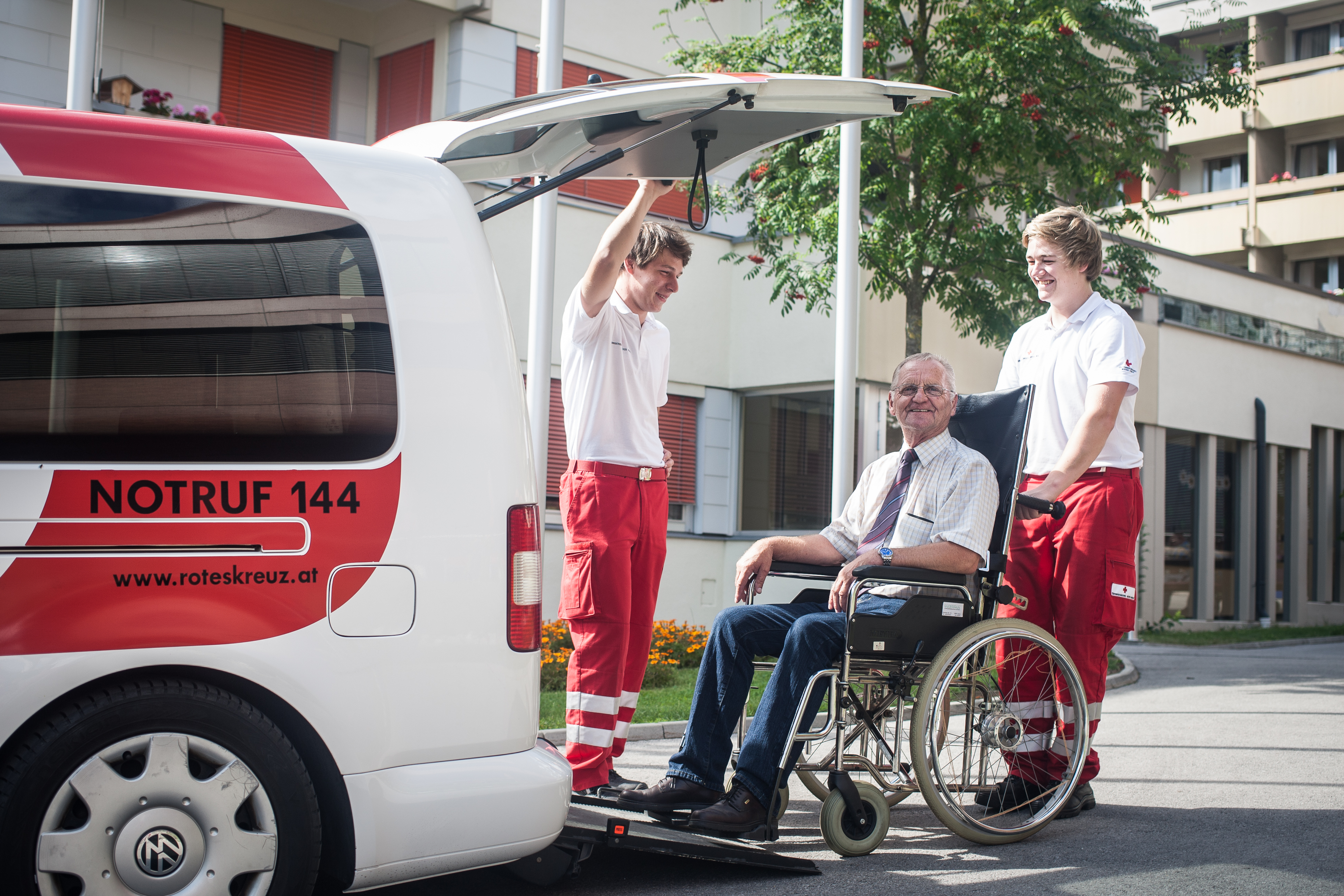 Zwei Sanitäter helfen einem Mann im Rollstuhl beim Einsteigen in ein Krankenwagen mit geöffnetem Heck. © ÖRK
