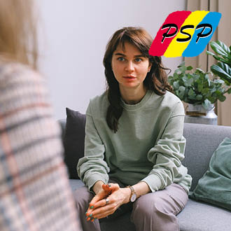 Eine junge Frau in einem grauen Pullover sitzt auf einem Sofa und hört aufmerksam zu. Grüne Pflanzen deuten auf eine entspannte Atmosphäre hin.