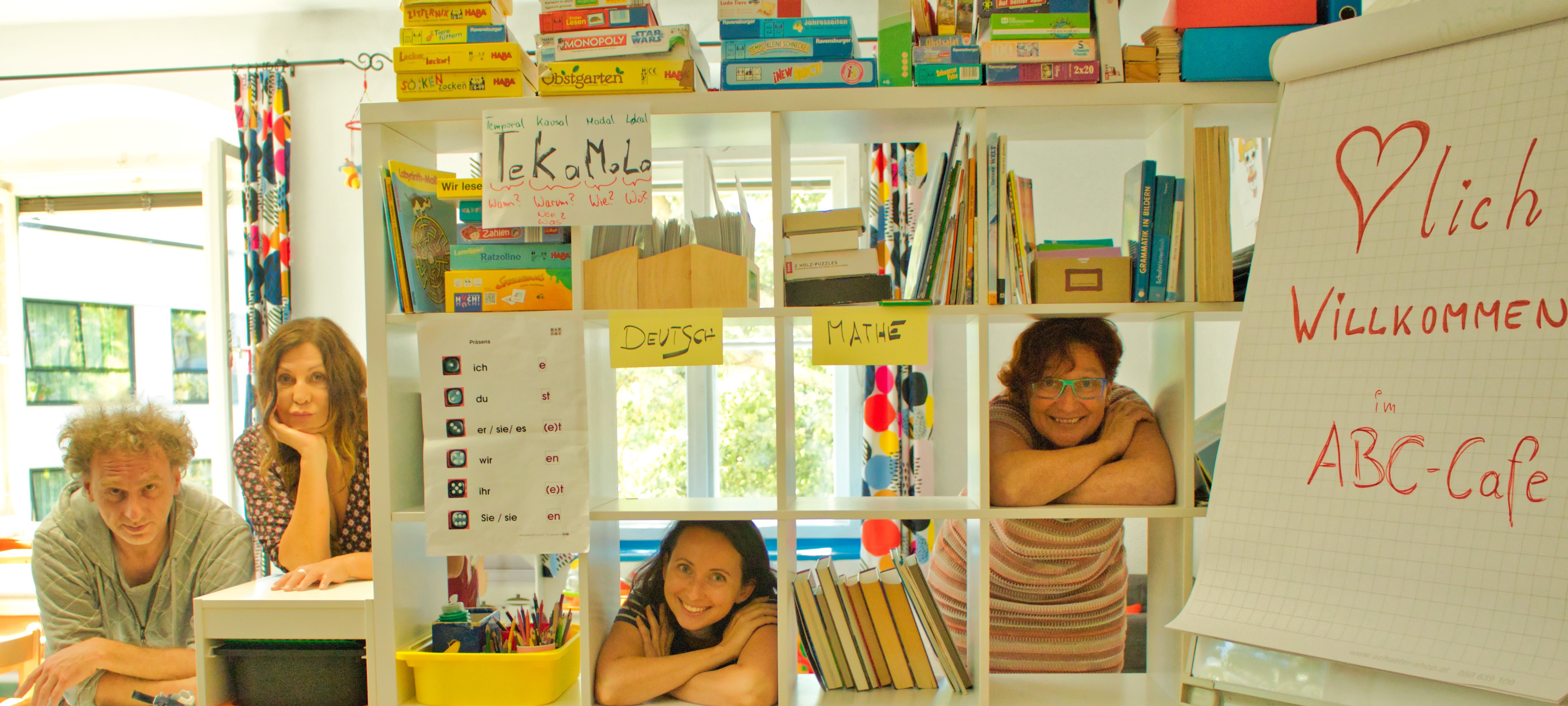 Vier Personen stehen hinter einem Bücherregal in einem freundlichen Raum mit Begrüßungsschildern für ein ABC-Café.