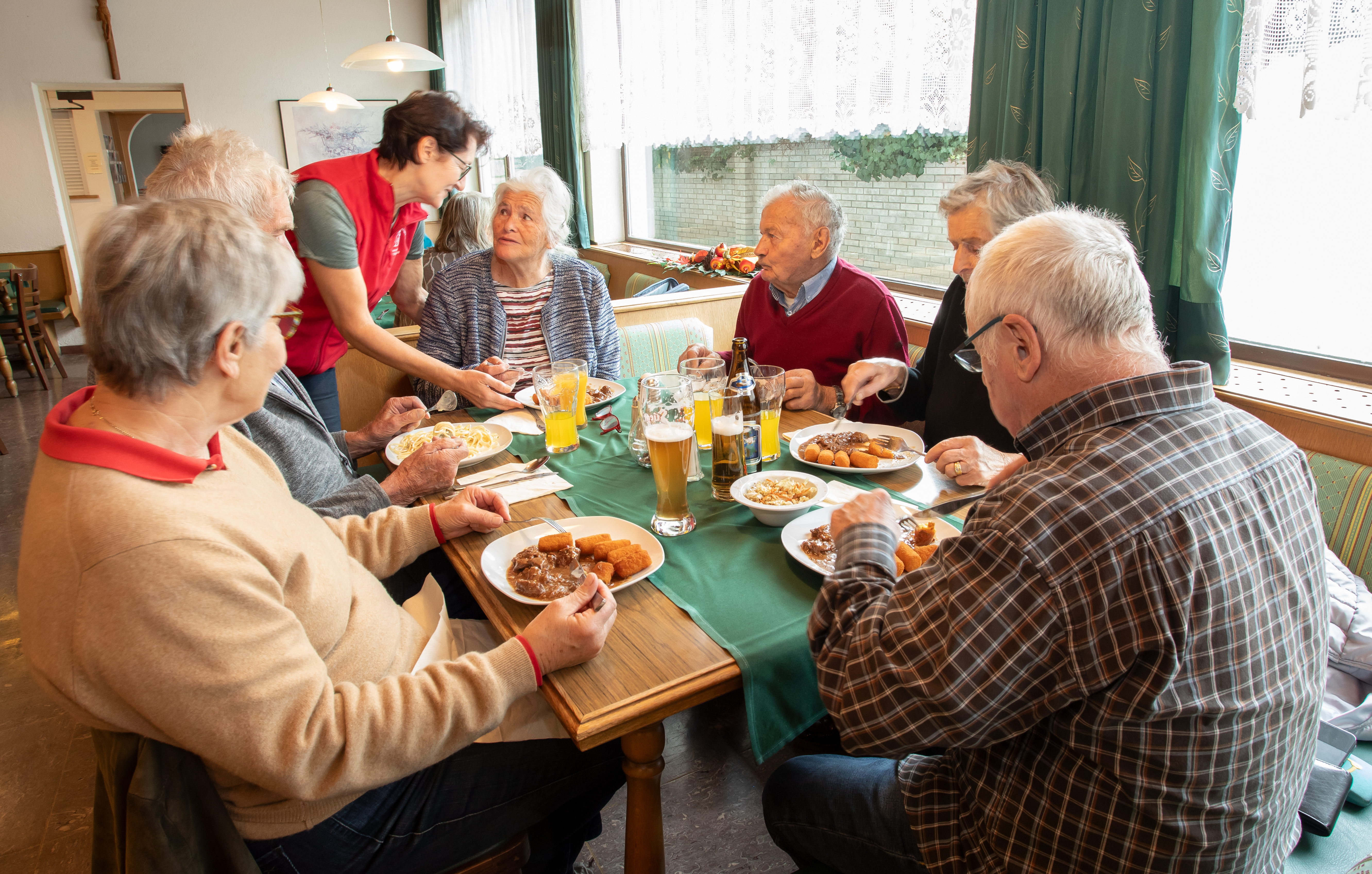 Eine Gruppe älterer Menschen sitzt an einem Tisch und genießt gemeinsam ein Essen und Getränke. Eine Bedienung serviert.