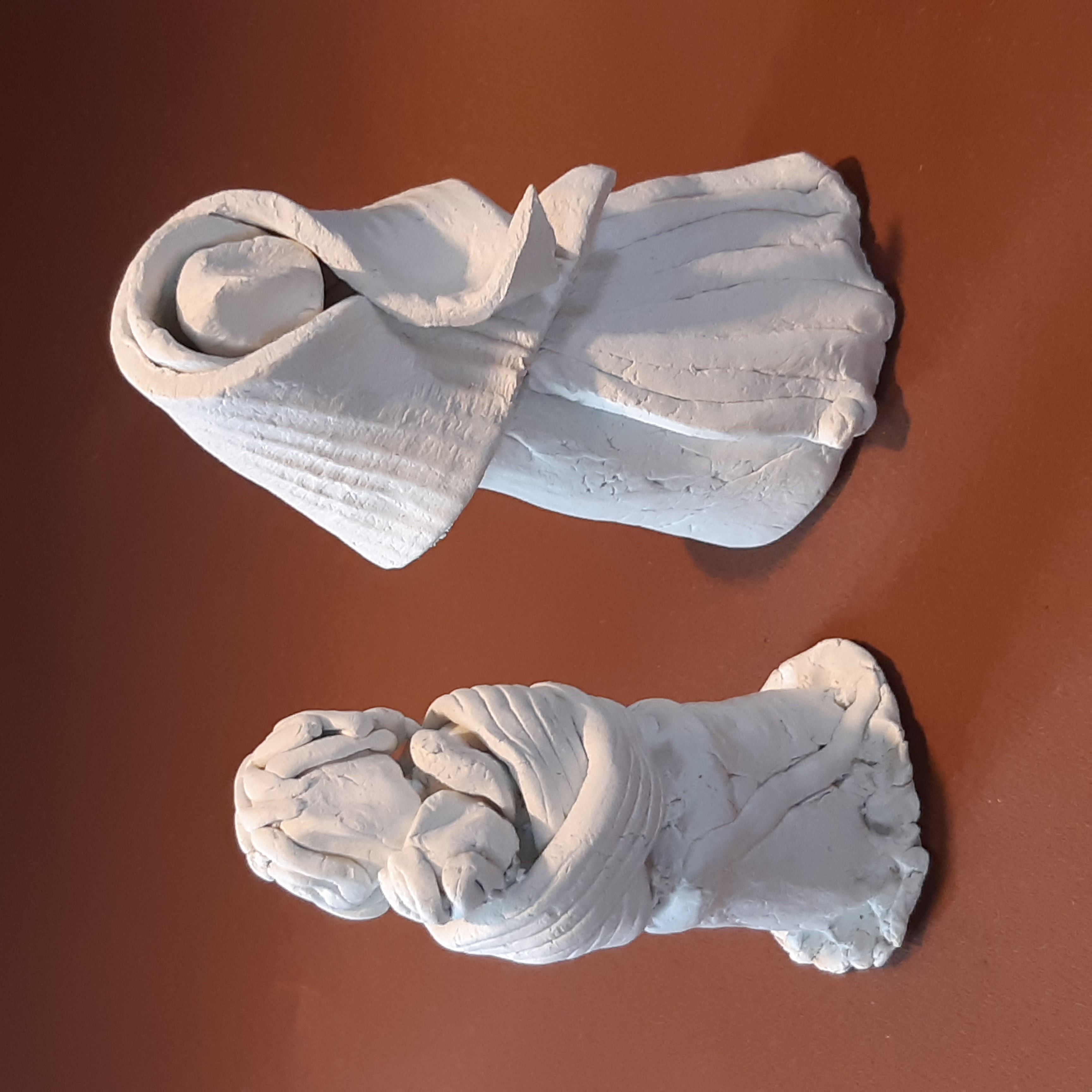 Zwei Figuren aus weißem Ton, eine umhüllte Statue und eine andere, die ein Kind hält, auf braunem Untergrund.