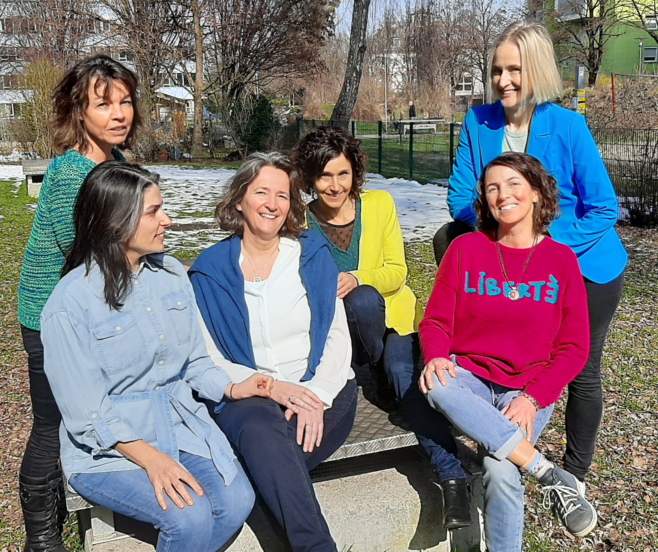Sechs Frauen sitzen auf einer Bank im Freien, lachen und genießen das sonnige Wetter in einem Park.