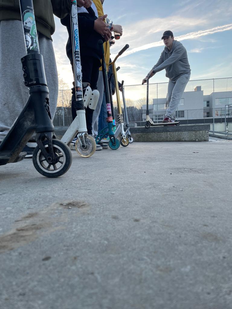 Gruppe von Personen mit Scootern, ein Fahrer springt von einer kleinen Plattform auf einem Skatepark.
